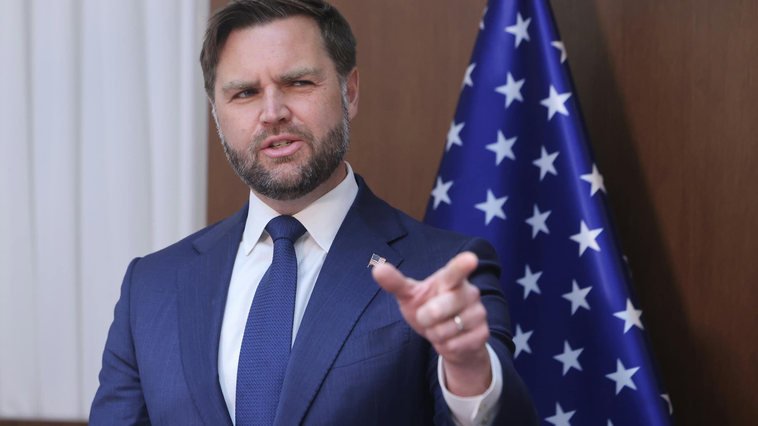 US vice-president, J.D. Vance points towards the camera while standing in front of a stars and stripes flag.