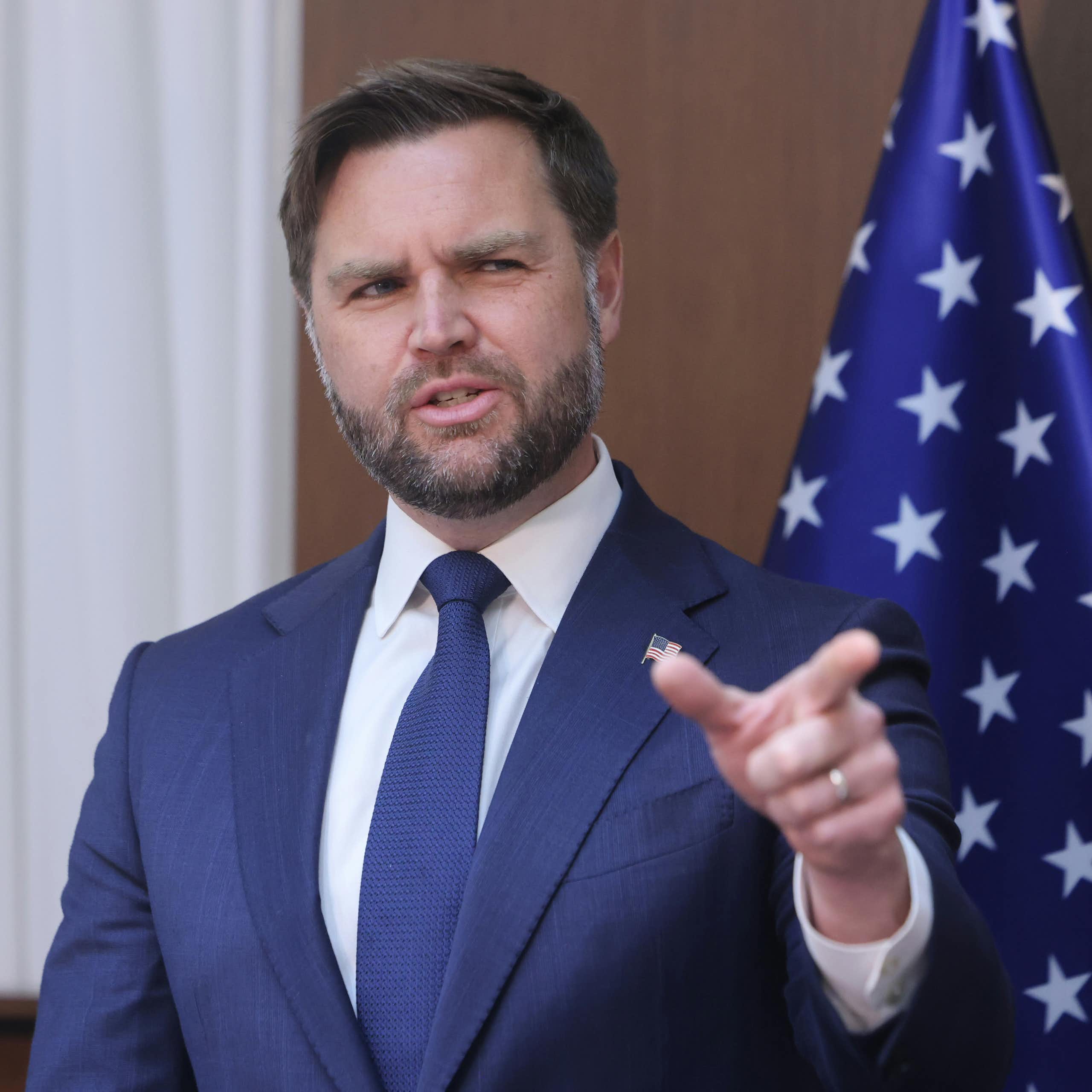 US vice-president, J.D. Vance points towards the camera while standing in front of a stars and stripes flag.