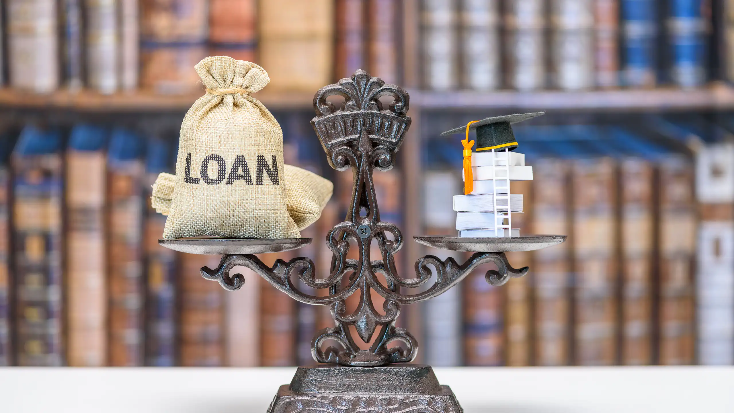 A metal scale shows a bag that says 'loan' on one side, with small books covered by a black graduation cap on the other side.