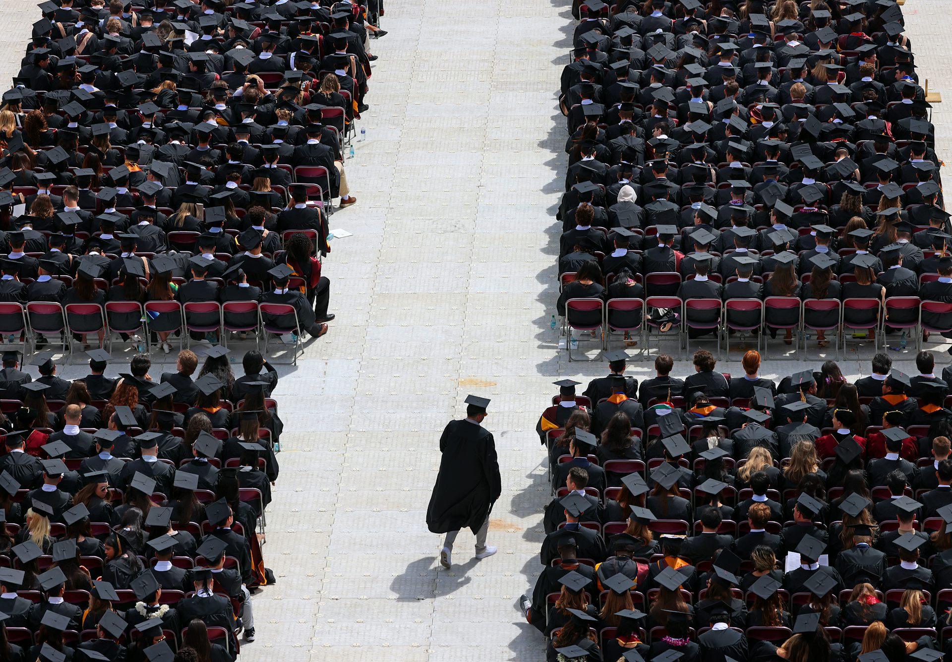 A large group of people dressed in black robes and graduation caps are seated in rows, except for one person who walks in the aisle.