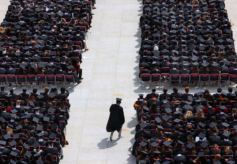 Trump is converting pupil mortgage forgiveness laws – barring some public staff from getting reduction, however resuming it for others 1 A large group of people dressed in black robes and graduation caps are seated in rows, except for one person who walks in the aisle.
