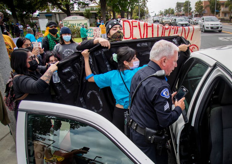 People wearing masks and holding signs surround a police car in front of a mobile home park.