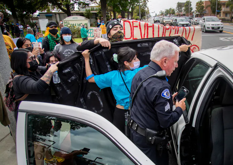 People wearing masks and holding signs surround a police car in front of a mobile home park.