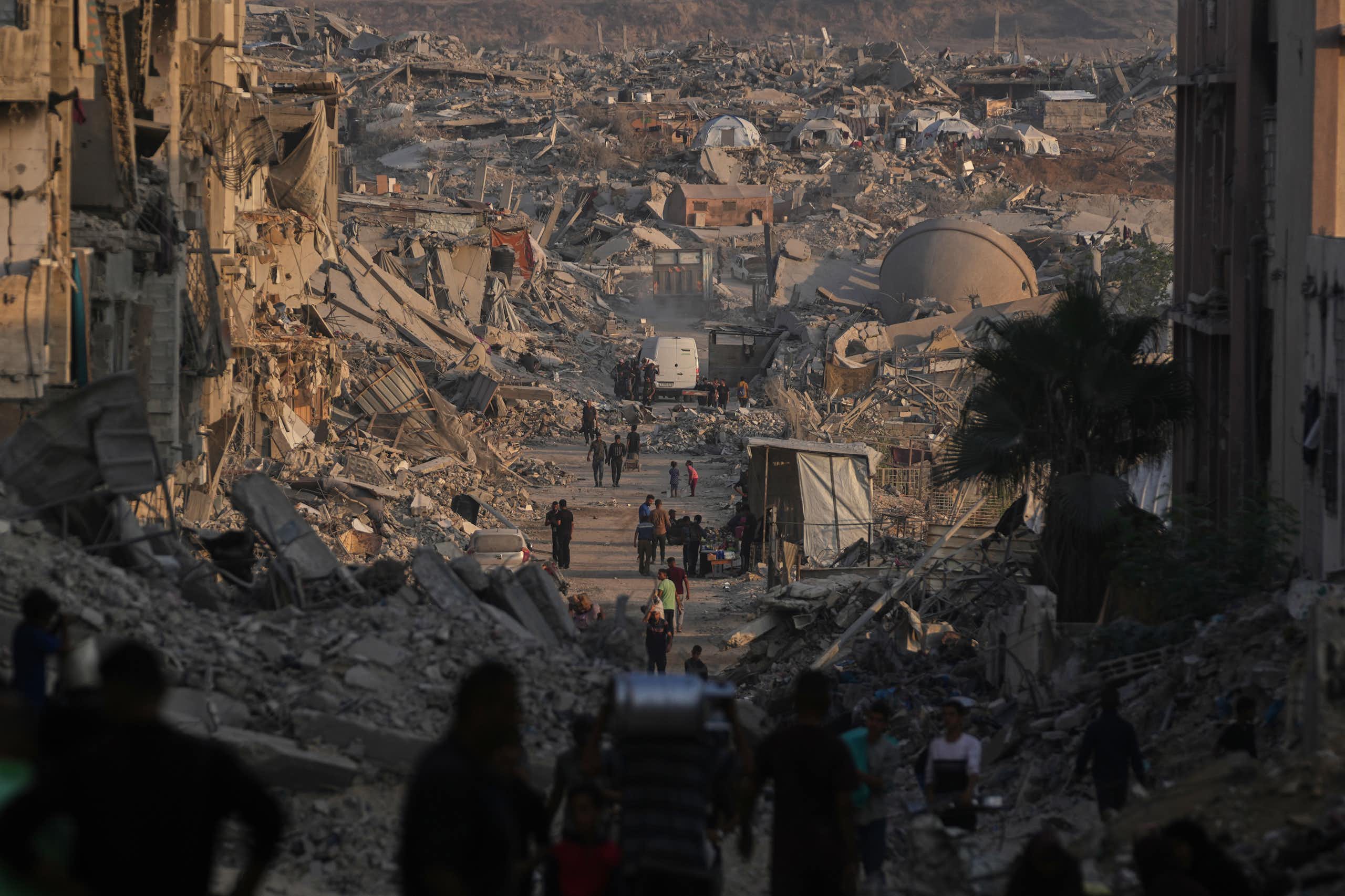 People walk along a road through a city that has been largely destroyed and left in rubble