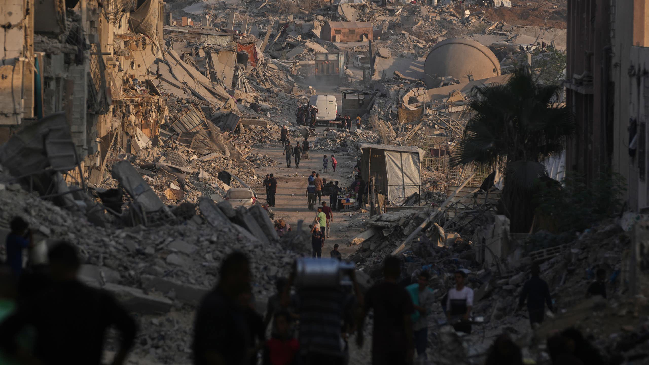 People walk along a road through a city that has been largely destroyed and left in rubble
