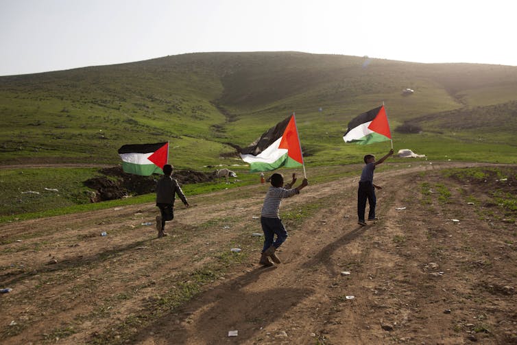 Tres niños corren por un camino de tierra, cada uno portando una bandera palestina.