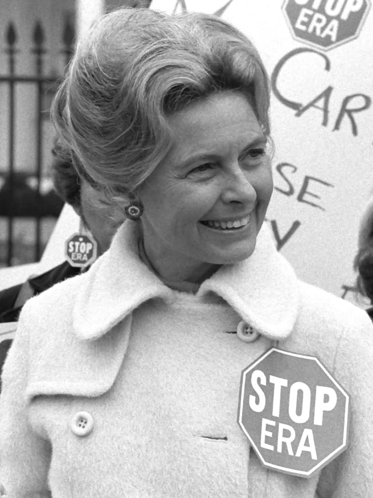 A black and white photo shows US conservative political activist Phyllis Schafly in a winter coat, and a badge fastened to it that reads 'stop ERA'. Her hair is done up and she is smiling at something out of view.