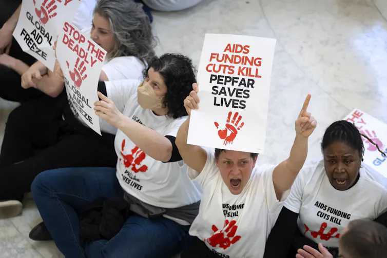 Protestors holding up signs and wearing white shirts reading 'AIDS FUNDING CUTS KILL PEPFAR SAVES LIVES' over a red handprint