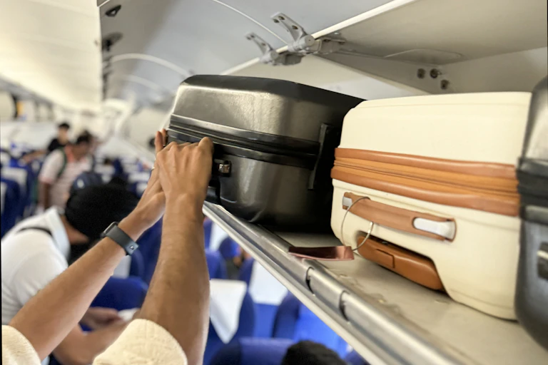 A close up of a man placing a grey wheelie suitcase in a full overhead baggage compartment in a plane.