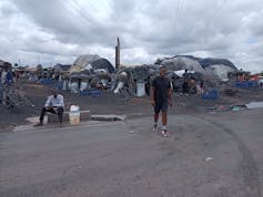 A man stands up in front of an area of home made plastic tents where people are selling small bags of home made charcoal