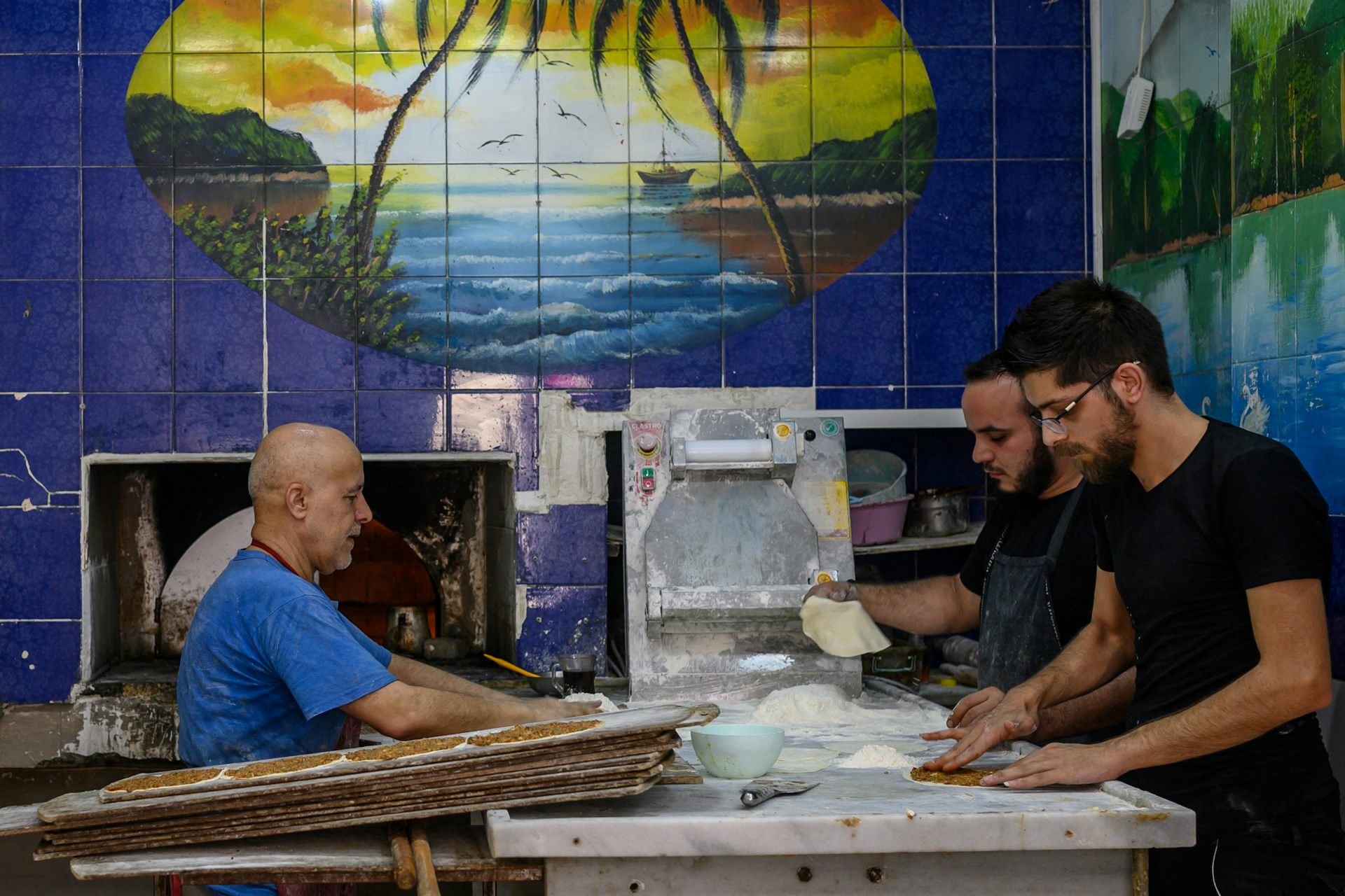 Three men work inside a bakery with blue-tiled walls.