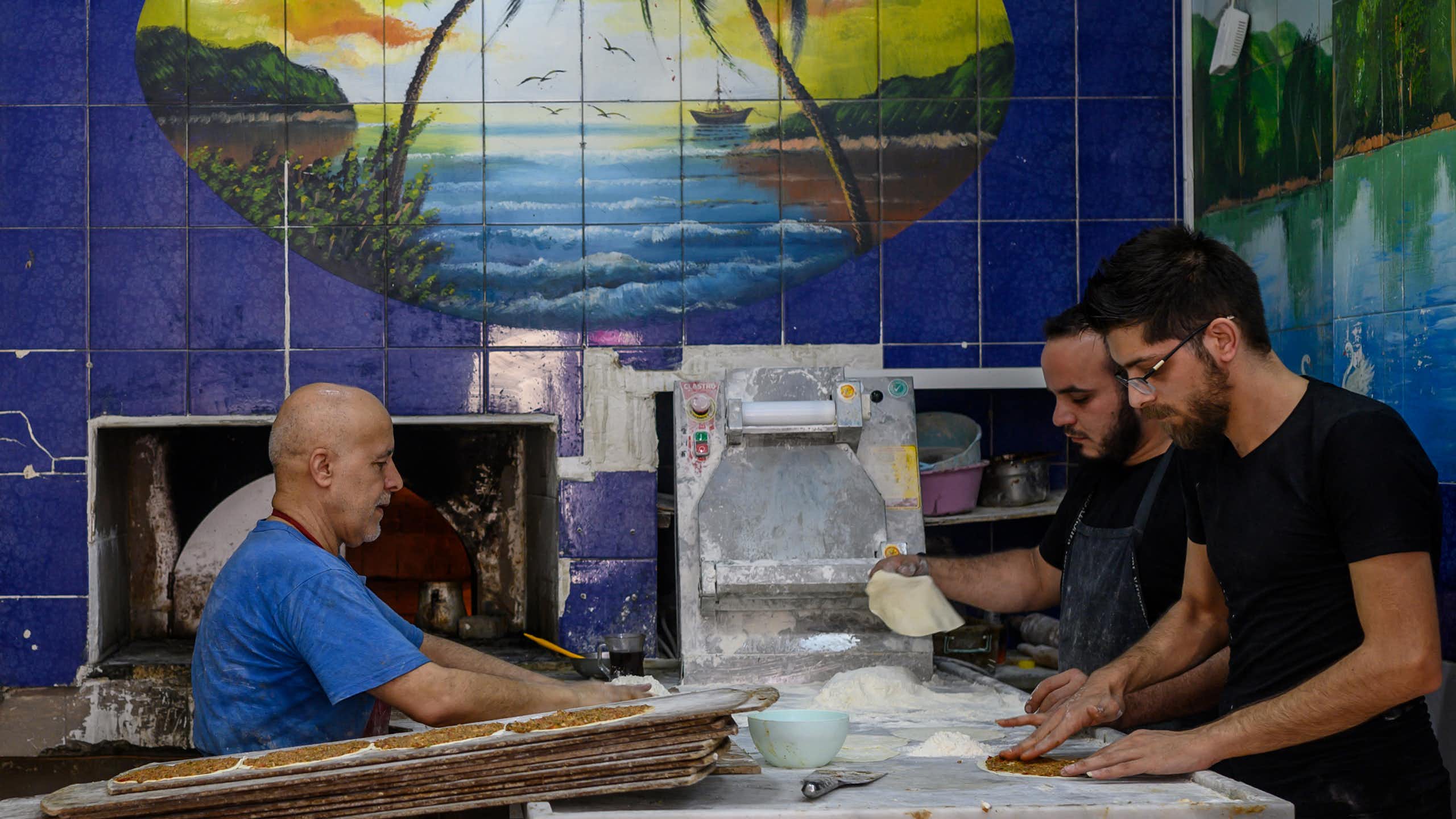 Three men work inside a bakery with blue-tiled walls.