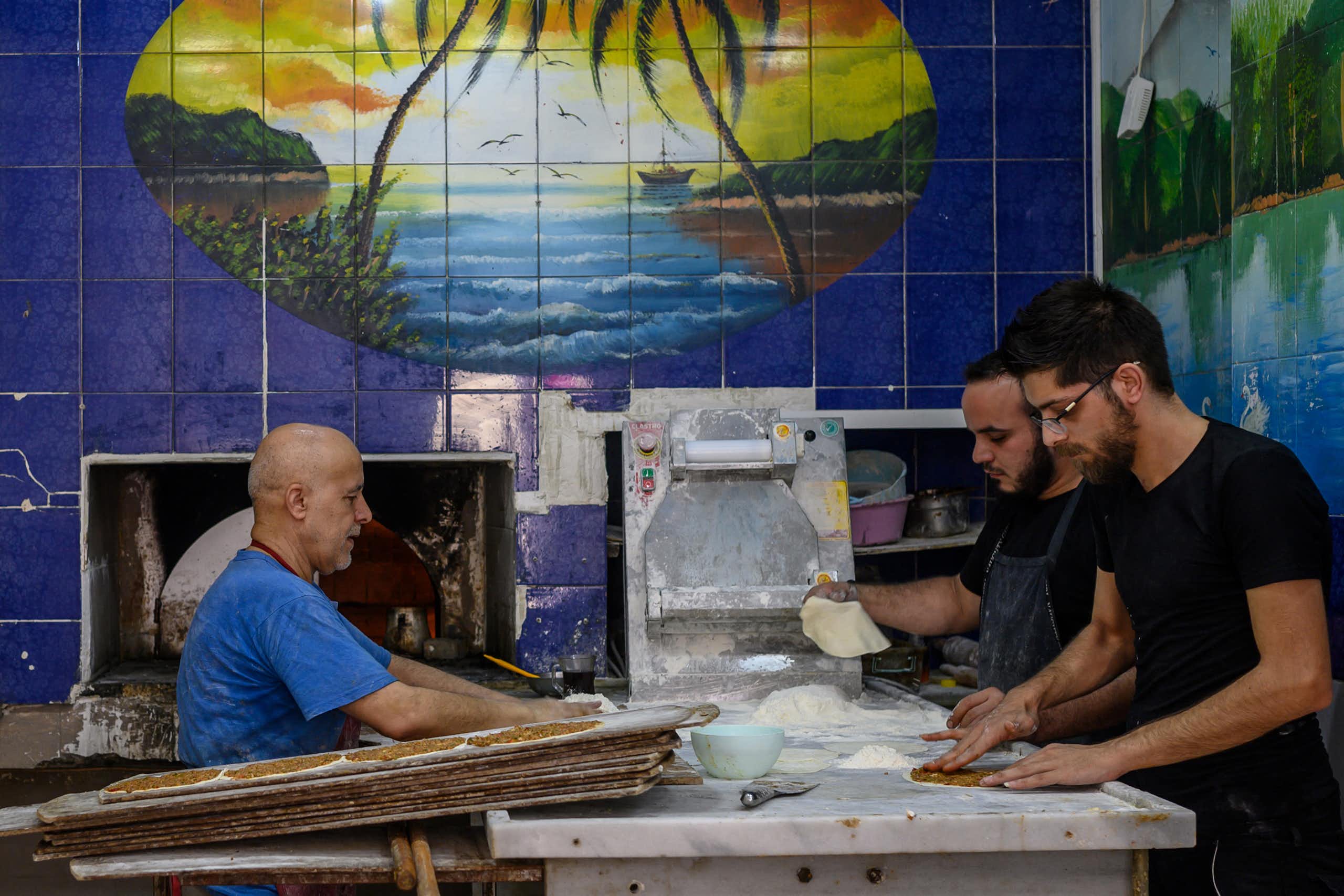 Three men work inside a bakery with blue-tiled walls.