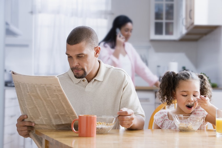 Dad reads newspaper at the kitchen table as toddler daughter has breakfast and wife stands in the kitchen in the background