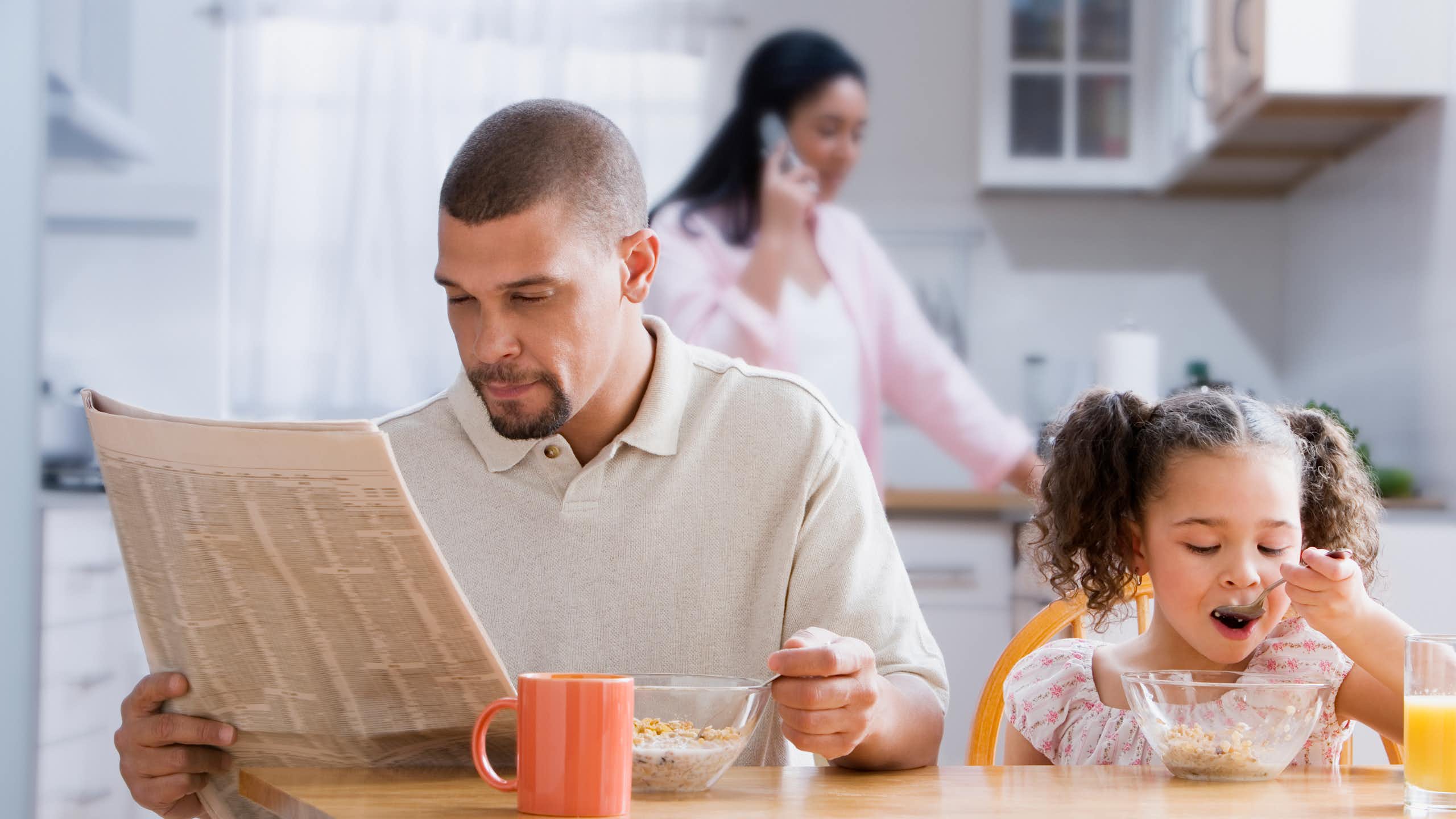Dad reads newspaper at the kitchen table as toddler daughter has breakfast and wife stands in the kitchen in the background