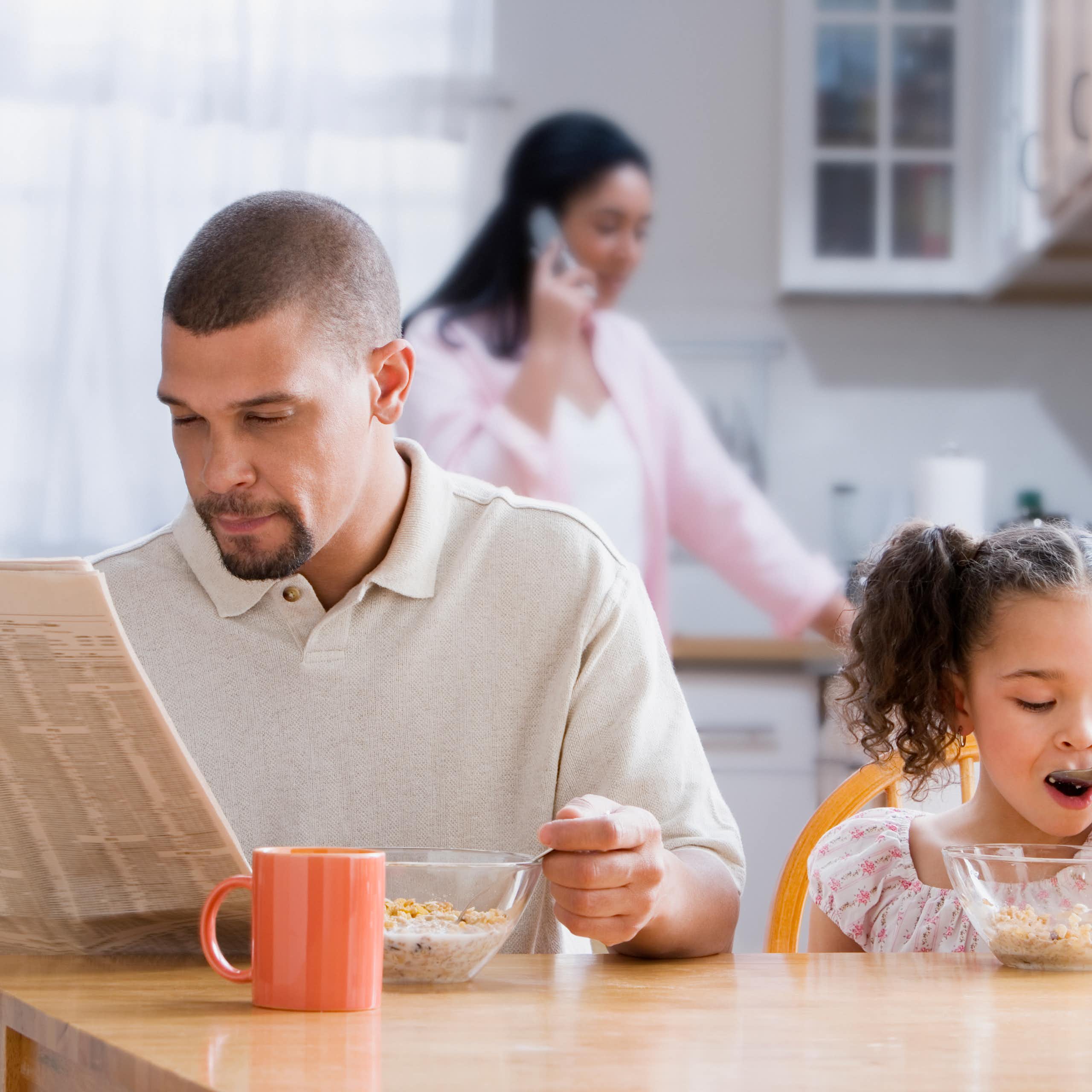 Dad reads newspaper at the kitchen table as toddler daughter has breakfast and wife stands in the kitchen in the background
