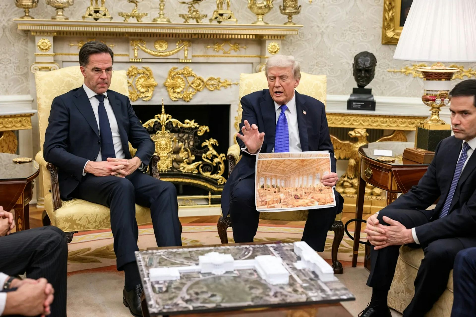 A man holds a photograph of a golden hall and explains something to other men sitting around.