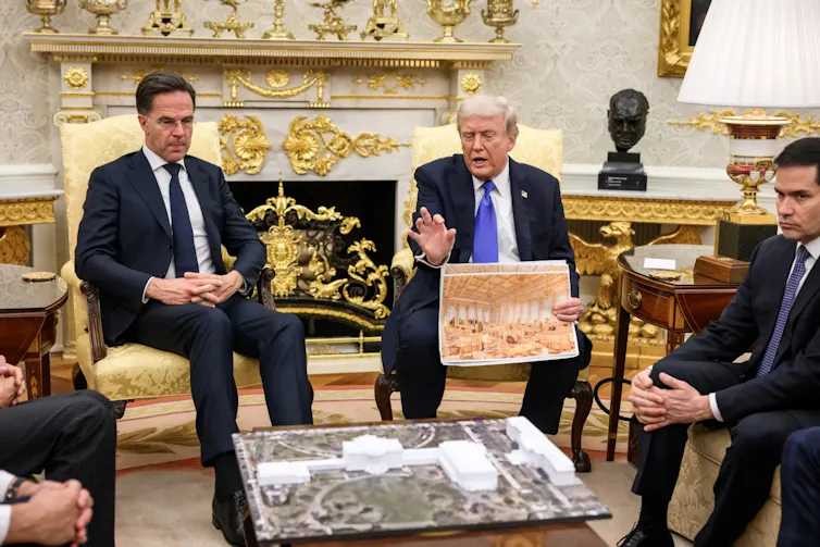 Un hombre sujeta una foto de un salón dorado y explica algo ante otros hombres sentados alrededor.