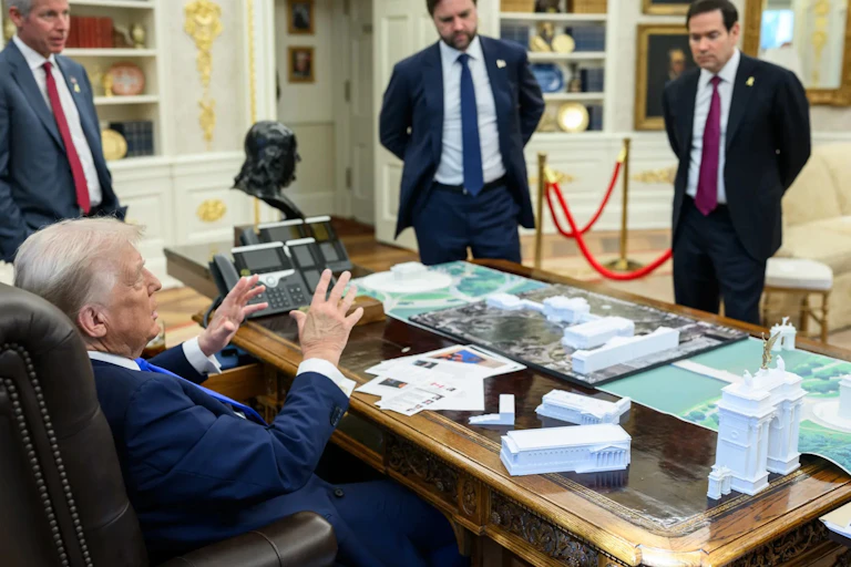 Un hombre habla desde una mesa con una maqueta con diferentes edificios sobre la mesa.