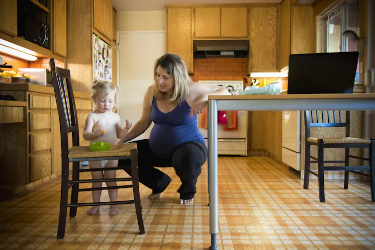 Pregnant mom squats near toddler in kitchen.