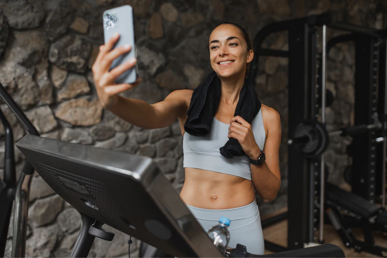 A smiling young woman livestreams from a treadmill at a luxury sports club.