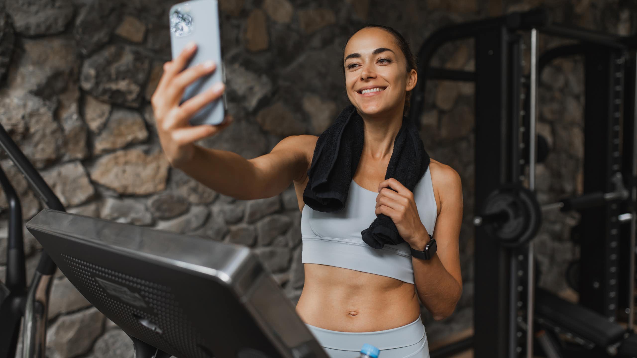 A smiling young woman livestreams from a treadmill at a luxury sports club.