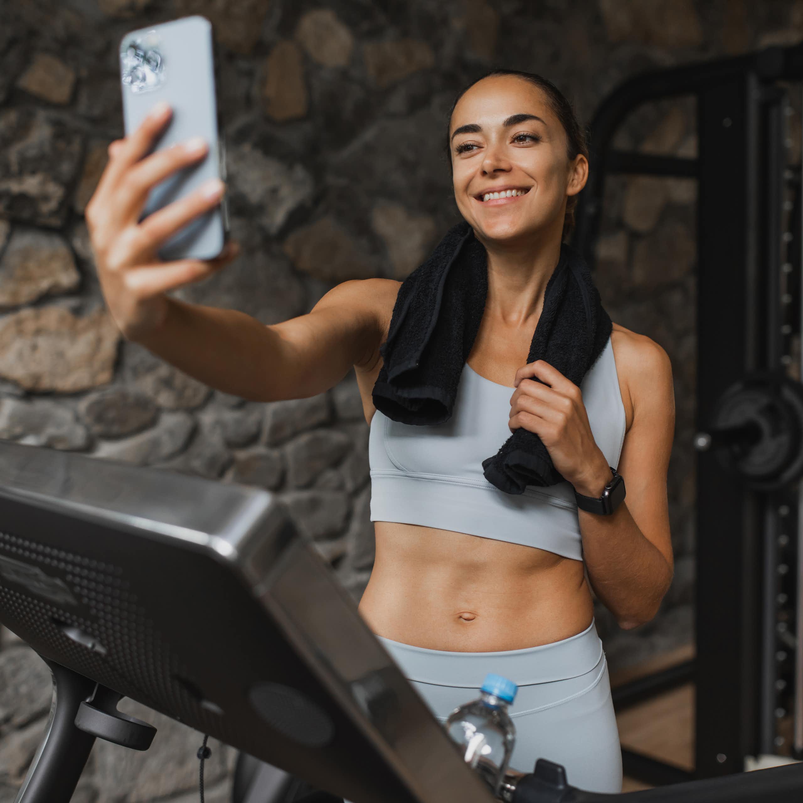 A smiling young woman livestreams from a treadmill at a luxury sports club.