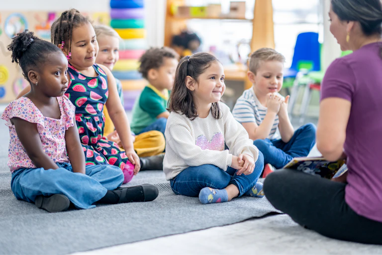 A group of kindergarten children sit on a rug while their teacher reads to them