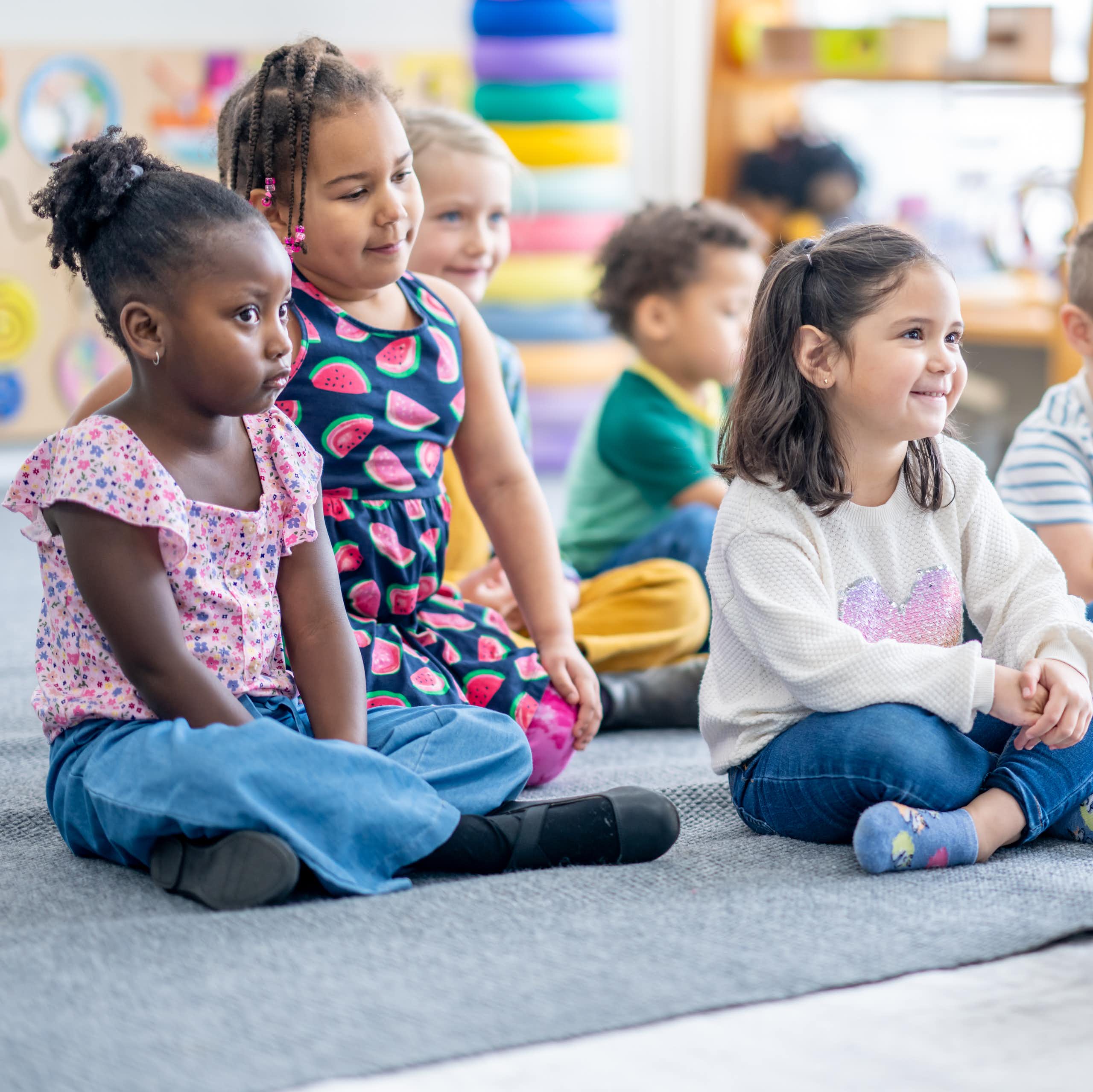 A group of kindergarten children sit on a rug while their teacher reads to them