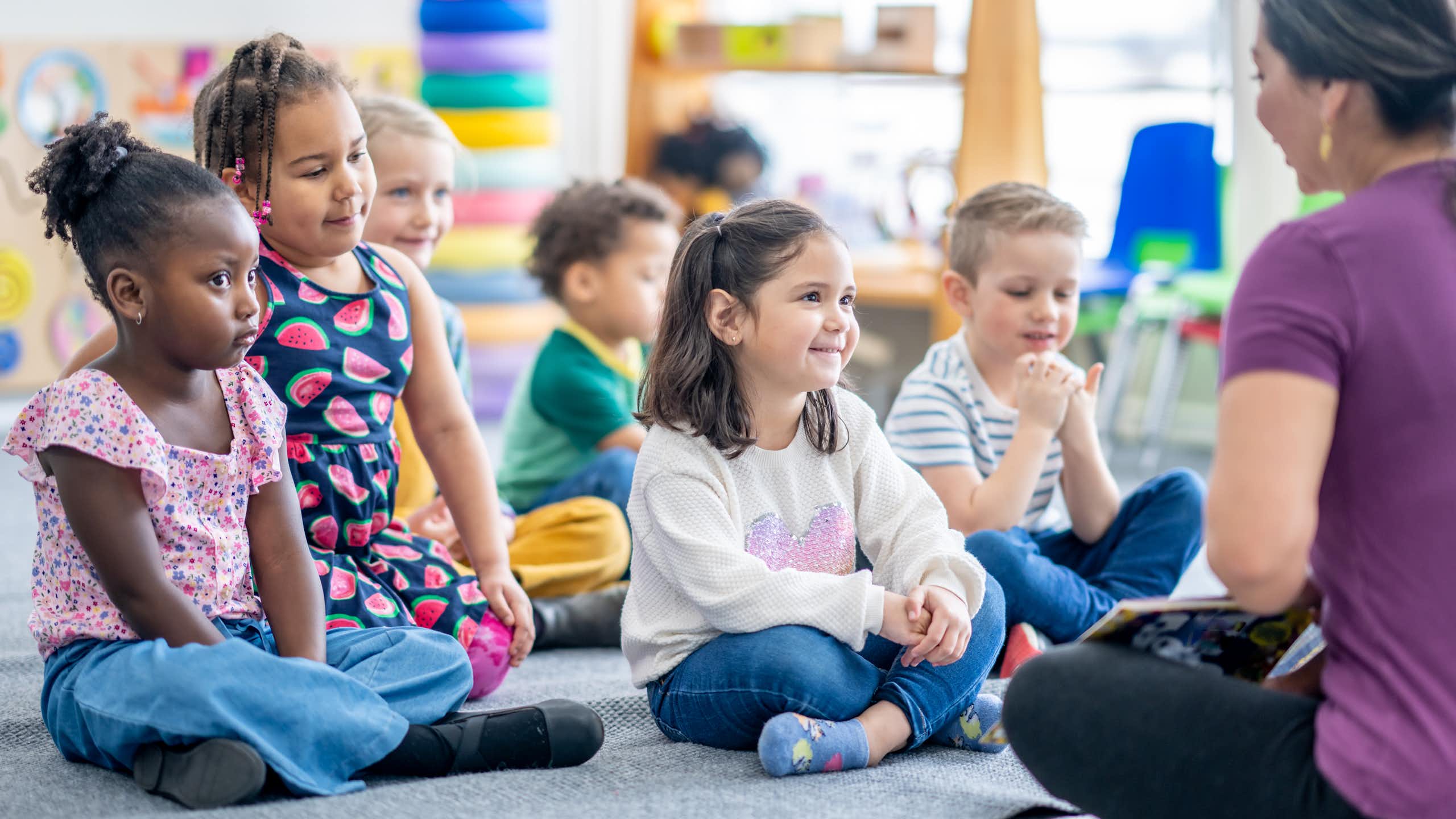 A group of kindergarten children sit on a rug while their teacher reads to them