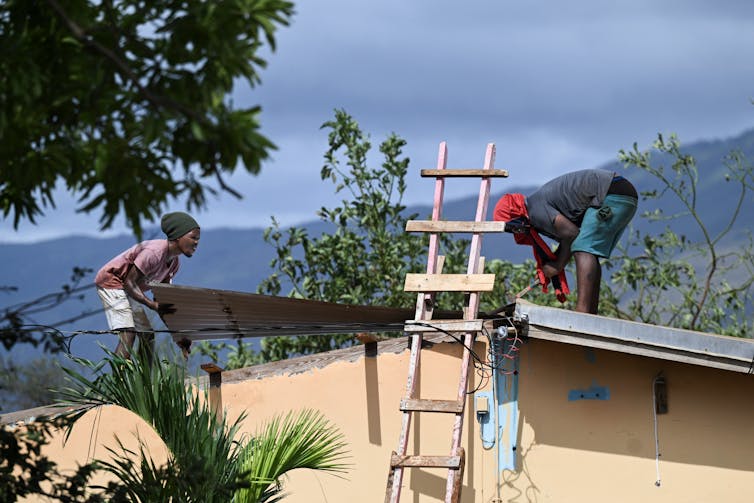 Two people put a piece of metal in place on a roof with a view of mountains in the background.