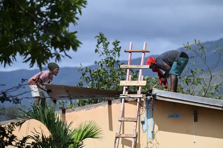 Two people put a piece of metal in place on a roof with a view of mountains in the background.