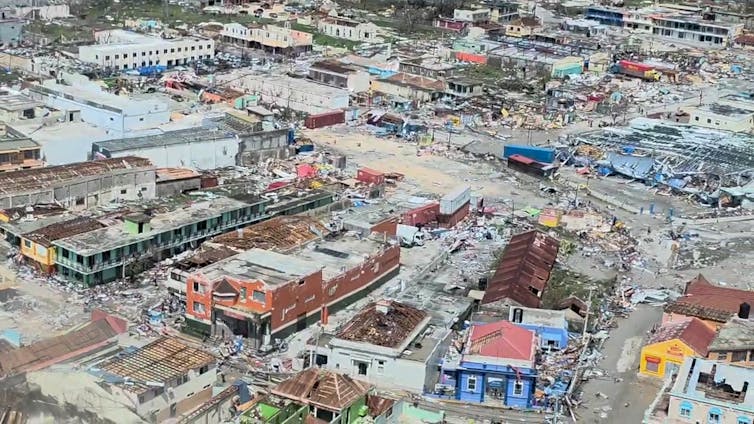 An aerial view of a business district shows buildings and homes with roofs and siding shredded, with mud covering the streets.
