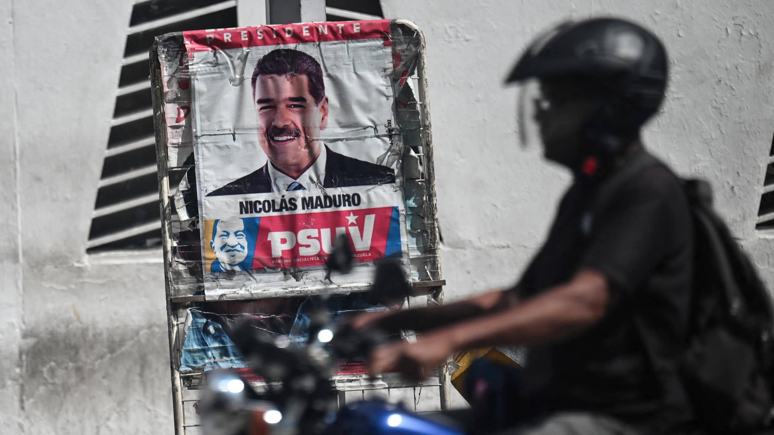 A man in a helmet and on a motorcycle drives past a poster with a a man's face.