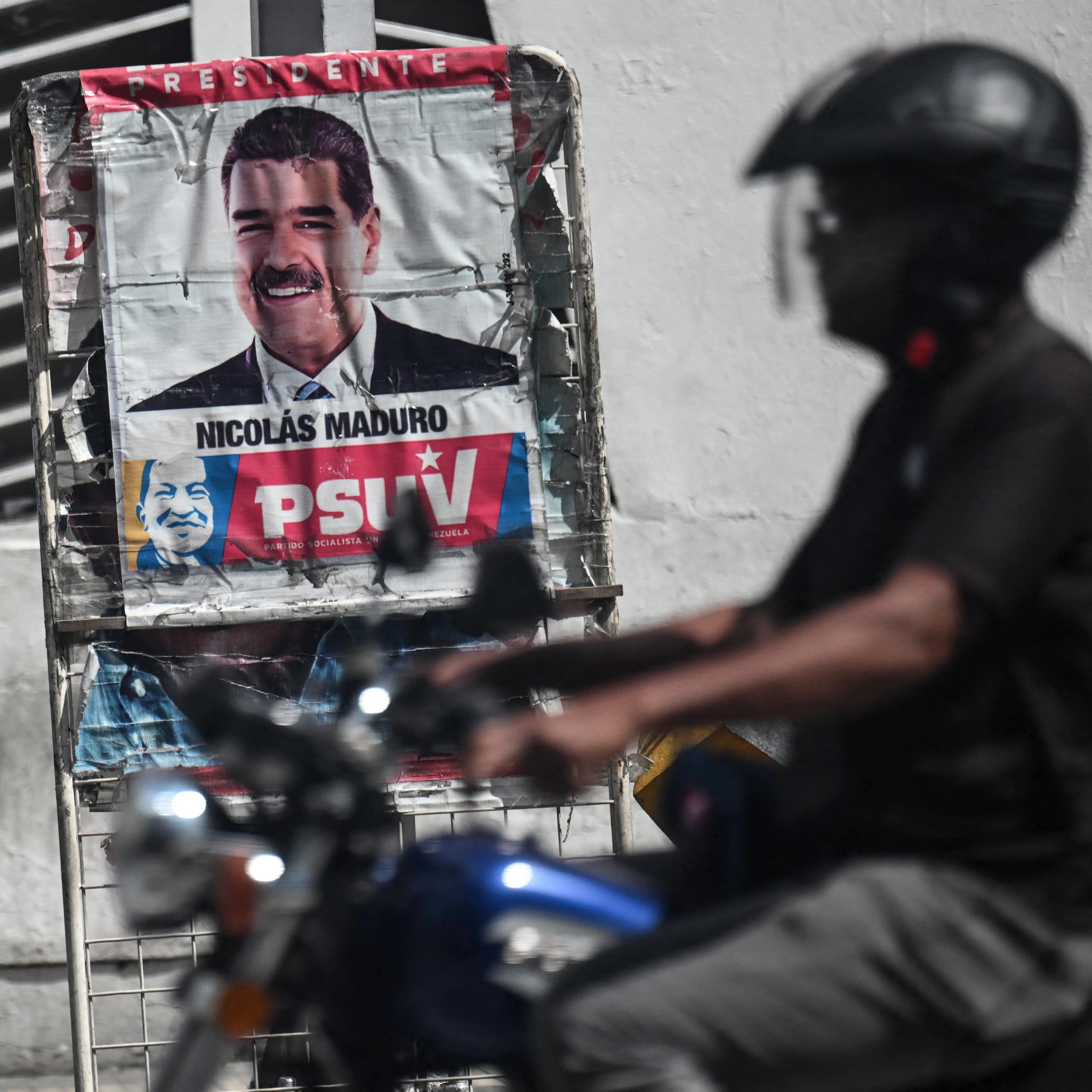 A man in a helmet and on a motorcycle drives past a poster with a a man's face.