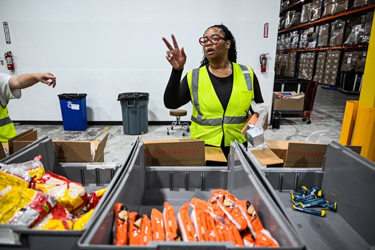 A worker gestures for more supplies while filling a cardboard box with package snacks.