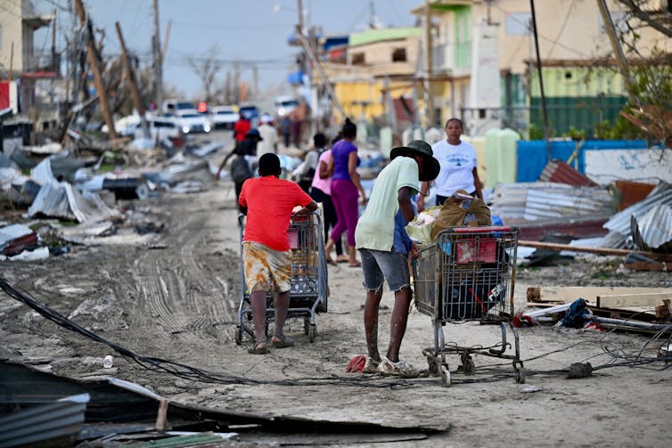 People push shopping carts on a muddy street with tangled power lines and damaged homes and vehicles.