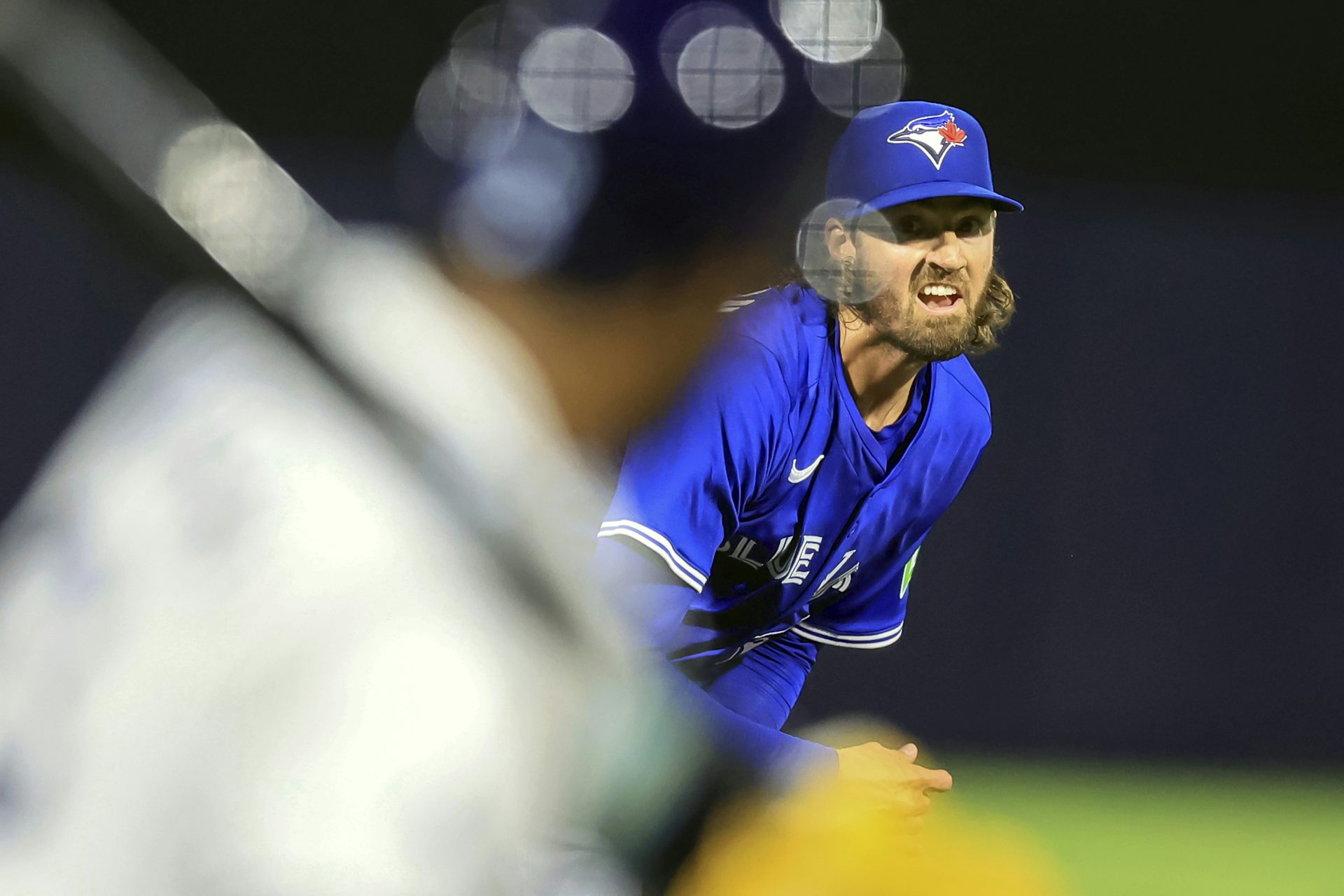A man in a dark blue Jays jersey, with a beard and longish hair, throws a pitch. the back of a batter is in the foreground.