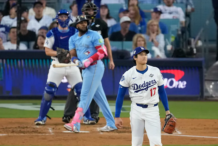 A Black man in a Blue Jays uniform looks up as an Asian pitcher in a Dodgers uniform looks in the same direction in dismay.