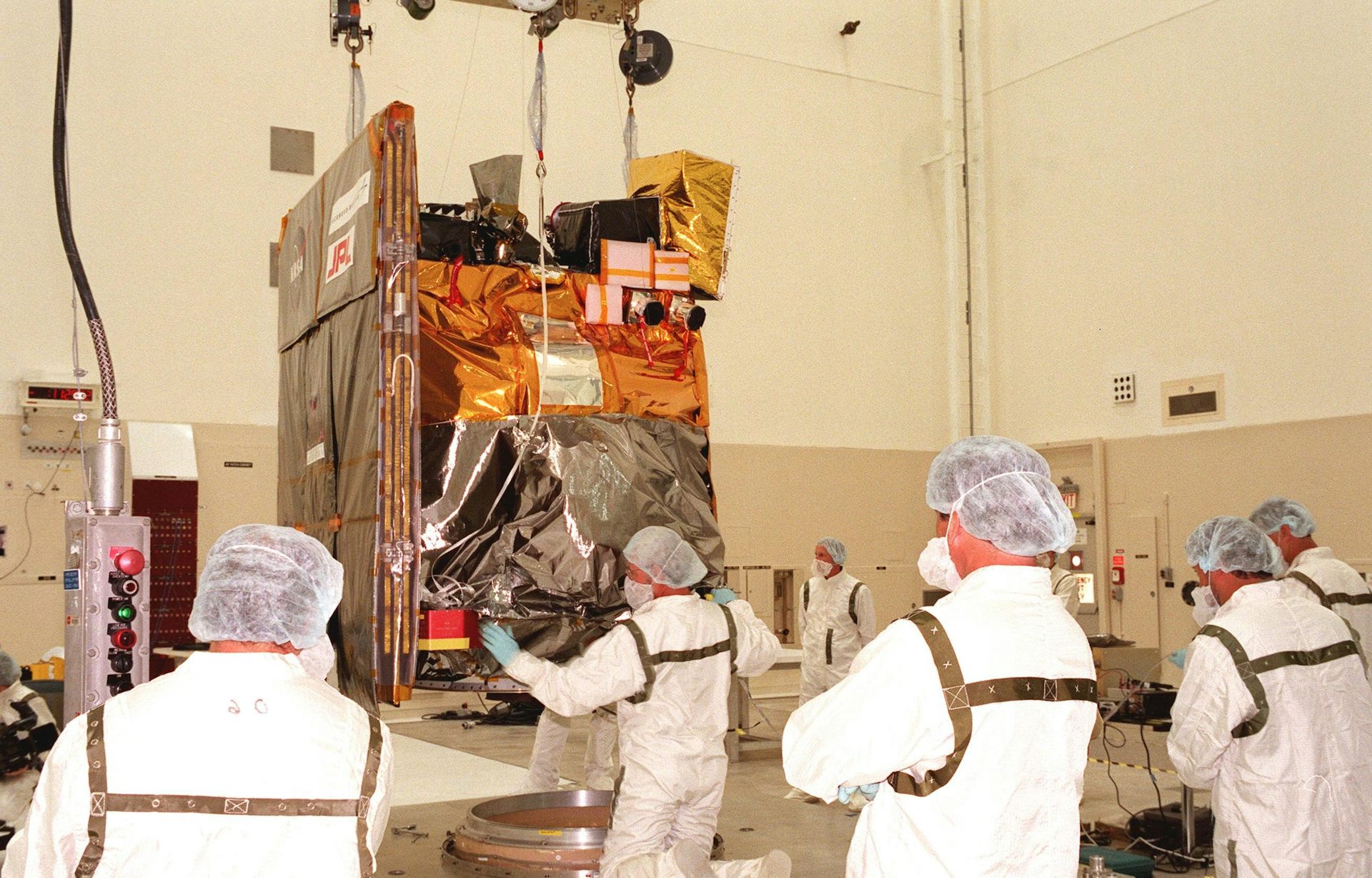 A group of people in white protective suits and head protectors stand in front of a robotic spacecraft