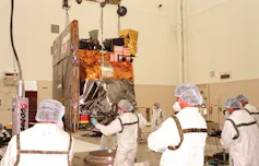 A group of people in white protective suits and head protectors stand in front of a robotic spacecraft