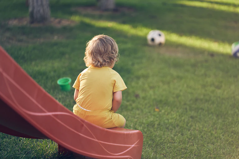 Child dressed in yellow sits at the end of a red slide, looking at a soccer ball on the grass