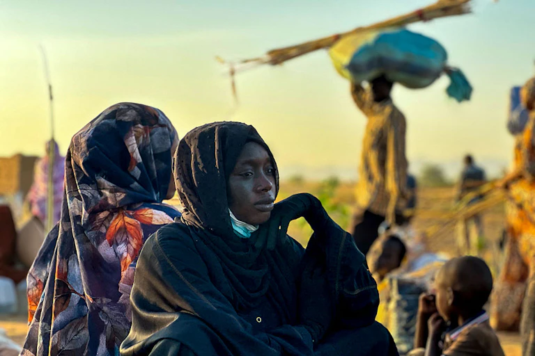 Women and children in the foreground sitting on the ground, with a man walking in the background with a sack on his head.