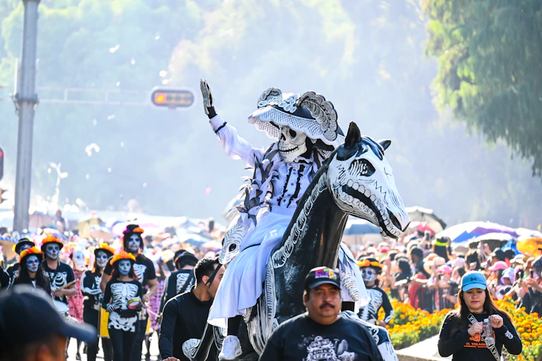 Una persona disfrazada de catrina saluda en un desfile con más gente disfrazada.