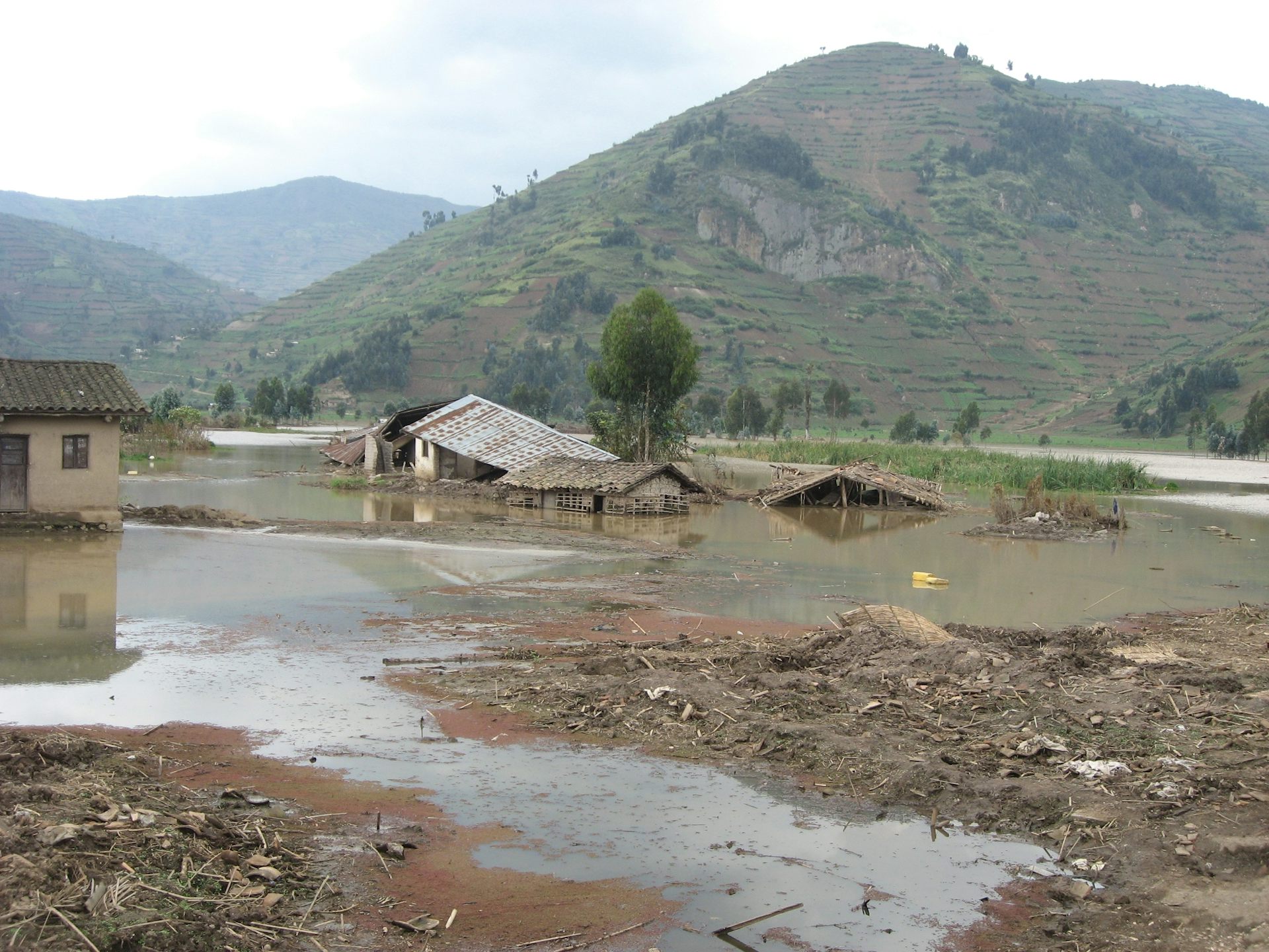 A flooded small farm with a mountain in the background, with buildings collapsing and crops covered with muddy water
