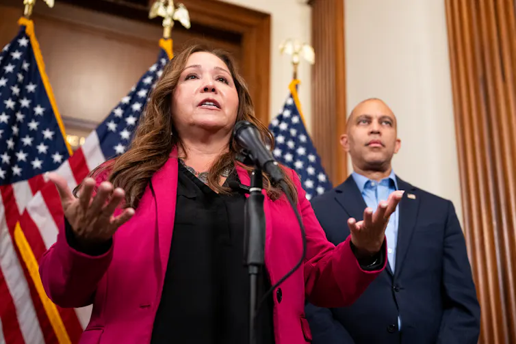 A brown-haired woman in a red jacket stands at a microphone in front of three American flags, speaking.