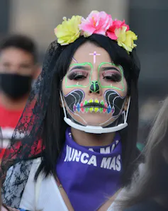 A young Mexican woman dressed as La Catrina with a neckerchief that says Nunca Mas (never again).