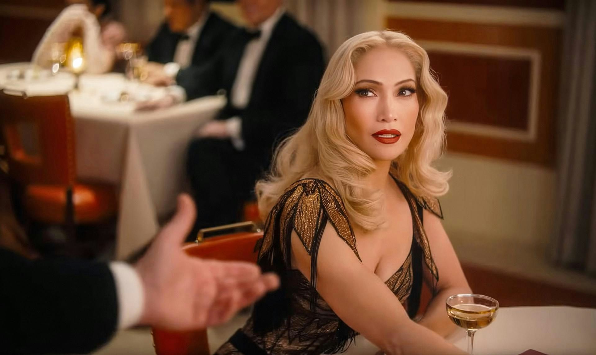 Una mujer sentada y vestida de gala observa a alguien que, fuera de cámara, le alarga la mano para invitarla a bailar.