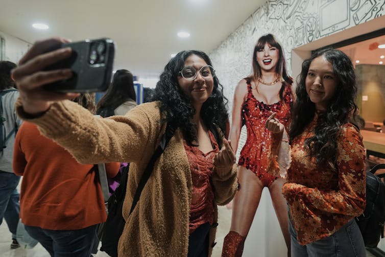 Two girls take a selfie with a cardboard Taylor Swift promo piece.