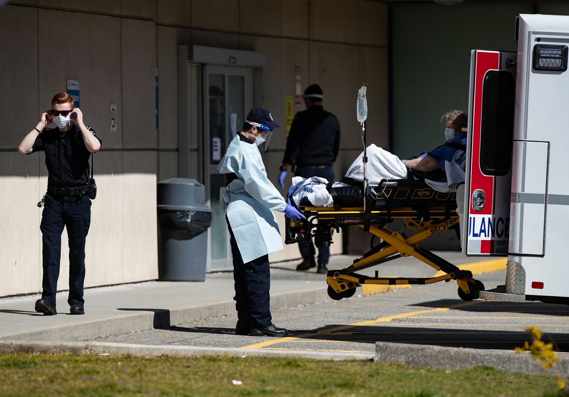 Un paramédico que lleva una mascarilla saca una camilla de una ambulancia mientras otro paramédico se pone una mascarilla en el fondo.
